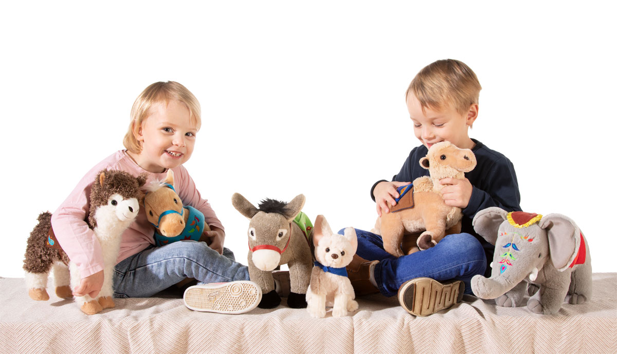 Zwei Kinder spielen mit Kuscheltieren auf einer Decke Zwei Kinder sitzen auf einer Decke und spielen mit verschiedenen Kuscheltieren wie einem Esel, einem Kamel und einem Elefanten.